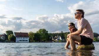 Two people are sitting on large rocks in the Rhine, cooling their feet in the water.