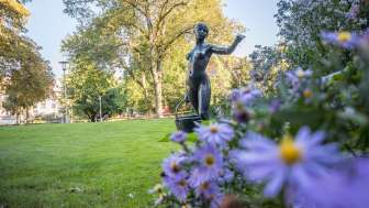 A statue of a woman carrying a basket in a park. There are blooming flowers in the foreground and trees in the background.