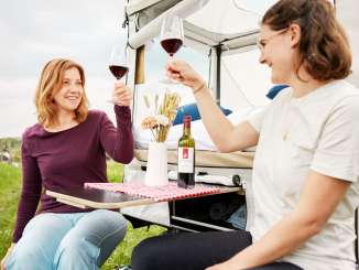 Two people toast with two full glasses of red wine. They are sitting in front of a camping tent.